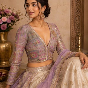 May include: A lavender long-sleeved blouse with a deep V-neckline and intricate multi-coloured embroidery, paired with a cream-coloured skirt featuring gold embellishments and a matching lavender sheer dupatta with a decorative gold border.