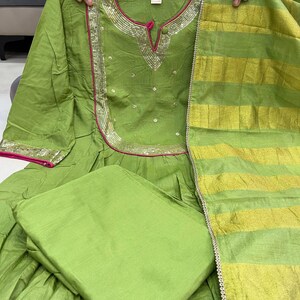 May include: A lime green Indian salwar kameez set. The top features a round neckline with silver and pink trim, and small embellishments. The matching dupatta has gold and green stripes. The set includes a solid green bottom.