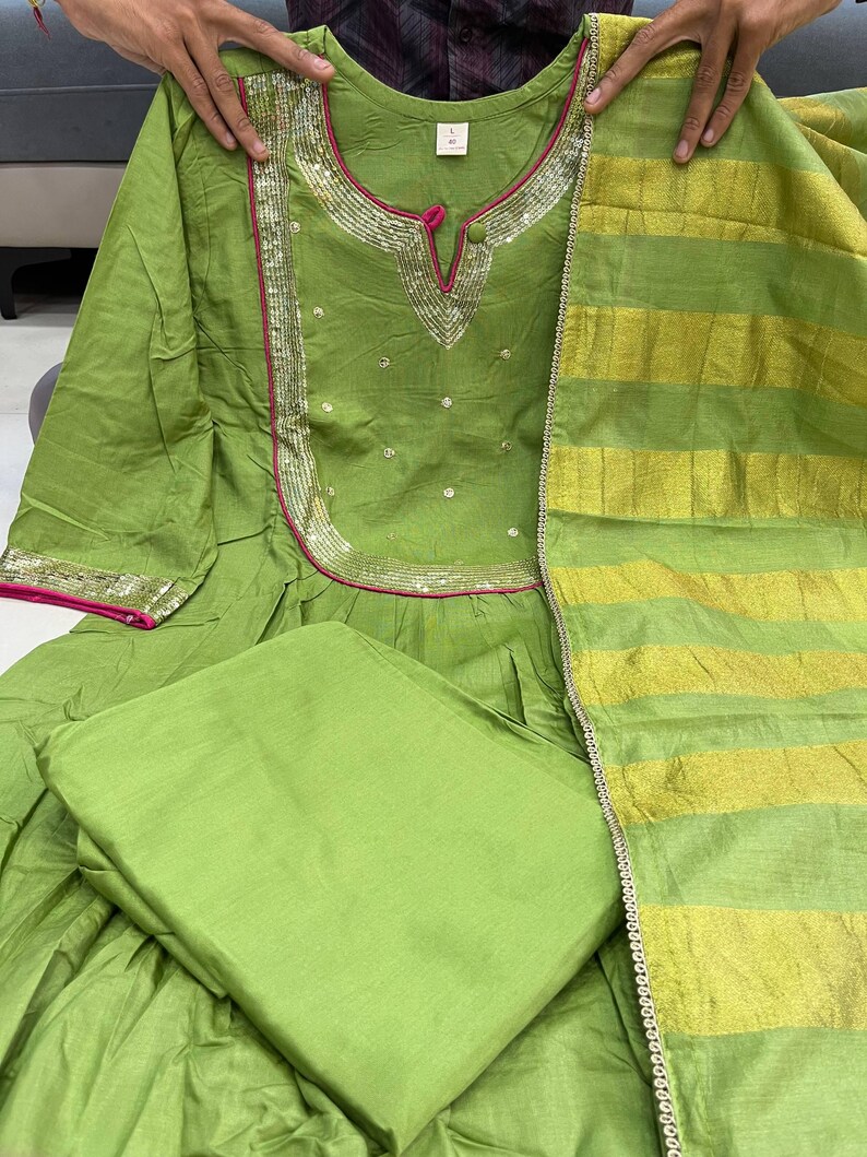 May include: A lime green Indian salwar kameez set. The top features a round neckline with silver and pink trim, and small embellishments. The matching dupatta has gold and green stripes. The set includes a solid green bottom.