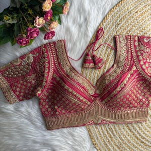 Peut inclure: Un chemisier rouge avec des broderies dorées complexes. Le chemisier présente un col en V profond et des manches courtes. La broderie comprend des motifs floraux et géométriques. Des glands pendent de l'encolure. Le chemisier fait partie d'une collection de vêtements traditionnels ou formels.