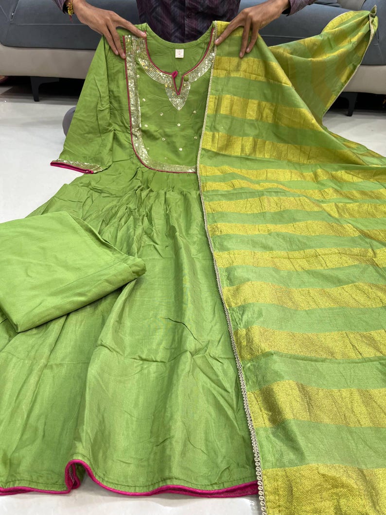 May include: A lime green Indian salwar kameez set. The dress features a round neckline with silver and gold embellishments, a pink trim, and a matching dupatta with gold stripes. The set includes a matching pair of pants.