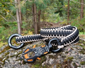 May include: Black and tan paracord lanyard with a black carabiner and an orange and black Frog 360 carabiner. The lanyard is made of woven cord and is designed for outdoor use. The carabiners are made of metal.