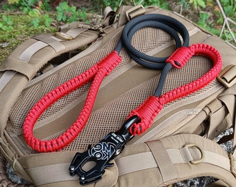 May include: A black and red paracord leash with a black metal clip, resting on a tan backpack. The leash features a braided red handle and a black and gray rope section. The clip has white text.