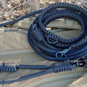 May include: A black paracord dog leash with a black metal clip. The leash is coiled and laid on a brown and tan fabric surface.