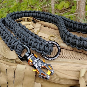 Op de afbeelding: Zwarte paracord-riem met een oranje en zwarte metalen clip. De riem is opgerold en ligt op een bruine rugzak.