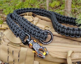 May include: Black paracord leash with an orange and black metal clip. The leash is coiled and resting on a tan backpack.