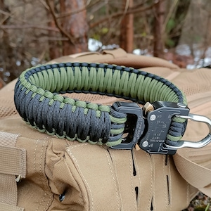 May include: A black and green paracord bracelet with a black buckle and a silver metal hook. The bracelet is lying on a tan fabric surface.