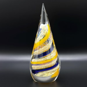 May include: A clear glass art piece in a teardrop shape, featuring yellow, blue, and gold swirling stripes. The top tapers to a sharp point. The decorative object is set against a black background.