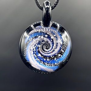 May include: A round, glass pendant with a swirling galaxy design. The pendant features shades of blue, white, and black, creating a cosmic effect. It hangs from a black cord, suitable for a necklace.