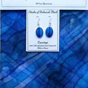 May include: A pair of blue silk earrings with silver plated metal components. The earrings are oval shaped and have a blue silk design. The earrings are made in Devon, England. The text on the card reads "Studio of Deborah Hack" and "Earrings 100% Silk with plated metal components Made in Devon."
