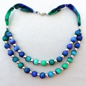 May include: A double-strand necklace with blue and green ombre beads separated by silver beads. The necklace is made with a green and blue patterned fabric.