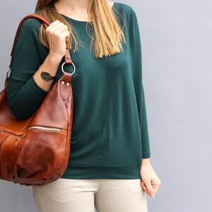May include: A brown leather shoulder bag with a zipper closure and a long strap. The bag is being held by a person wearing a teal green sweater and beige pants.