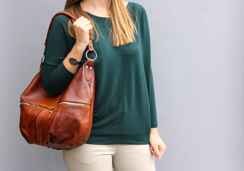 May include: A brown leather shoulder bag with a zipper closure and a long strap. The bag is being held by a person wearing a teal green sweater and beige pants.