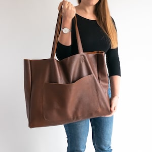 Könnte beinhalten: Eine große braune Ledertasche mit einer Tasche auf der Vorderseite. Die Tasche hat eine große Öffnung und ist aus weichem, strukturiertem Leder gefertigt.