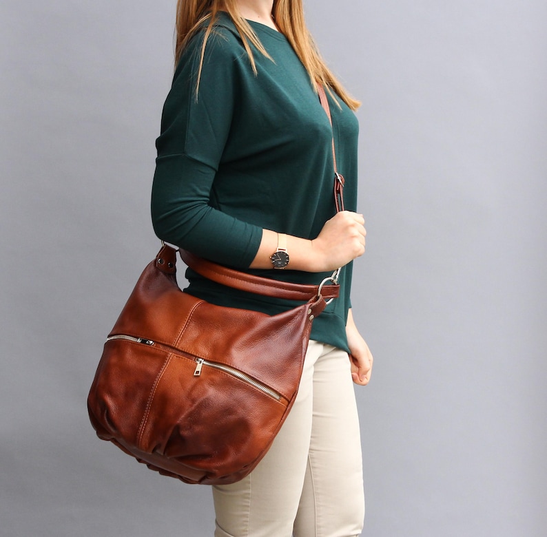 May include: A brown leather shoulder bag with a zippered pocket and a long strap. The bag is worn by a person wearing a green sweater and beige pants.
