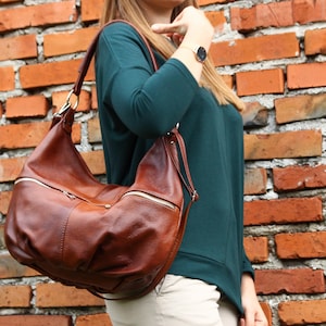 May include: A brown leather satchel bag with a zippered pocket and a long strap. The bag is worn over the shoulder of a person wearing a teal shirt.