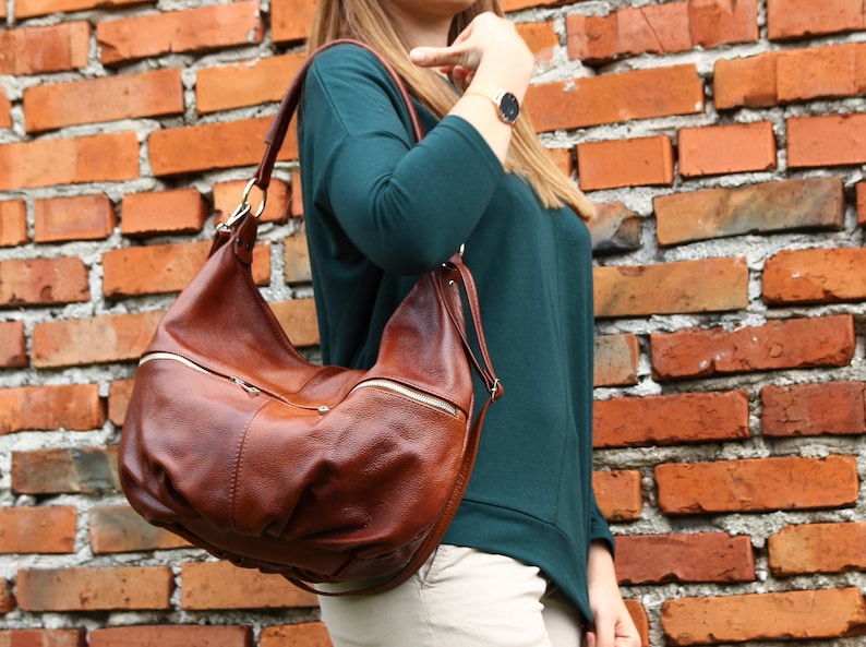 May include: A brown leather satchel bag with a zippered pocket and a long strap. The bag is worn over the shoulder of a person wearing a teal shirt.