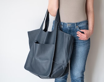 May include: A large, dark blue leather tote bag with a pebbled texture. The bag features two long shoulder straps and a prominent, deep exterior pocket on the front. The design is unstructured and slouchy, showcasing a deep navy hue.