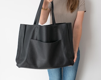 May include: A large black leather tote bag with a front pocket. The bag has a textured finish and is being held by a person.
