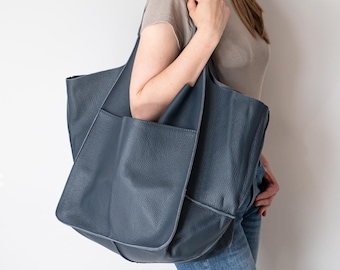 May include: A large, dark blue leather tote bag with a pebbled texture. The bag features a prominent, wide exterior pocket on the front and two sturdy shoulder straps. The design is spacious and unstructured, showcasing a deep blue hue.