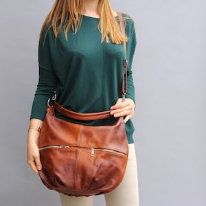 May include: A brown leather shoulder bag with a zipper closure and a long strap. The bag is worn by a person wearing a green sweater and beige pants.