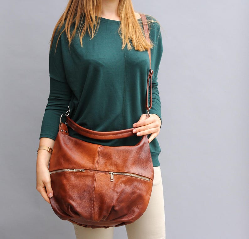 May include: A brown leather shoulder bag with a zipper closure and a long strap. The bag is worn by a person wearing a green sweater and beige pants.