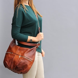 May include: A brown leather shoulder bag with a zippered pocket and a long strap. The bag is worn by a person wearing a teal sweater and beige pants.