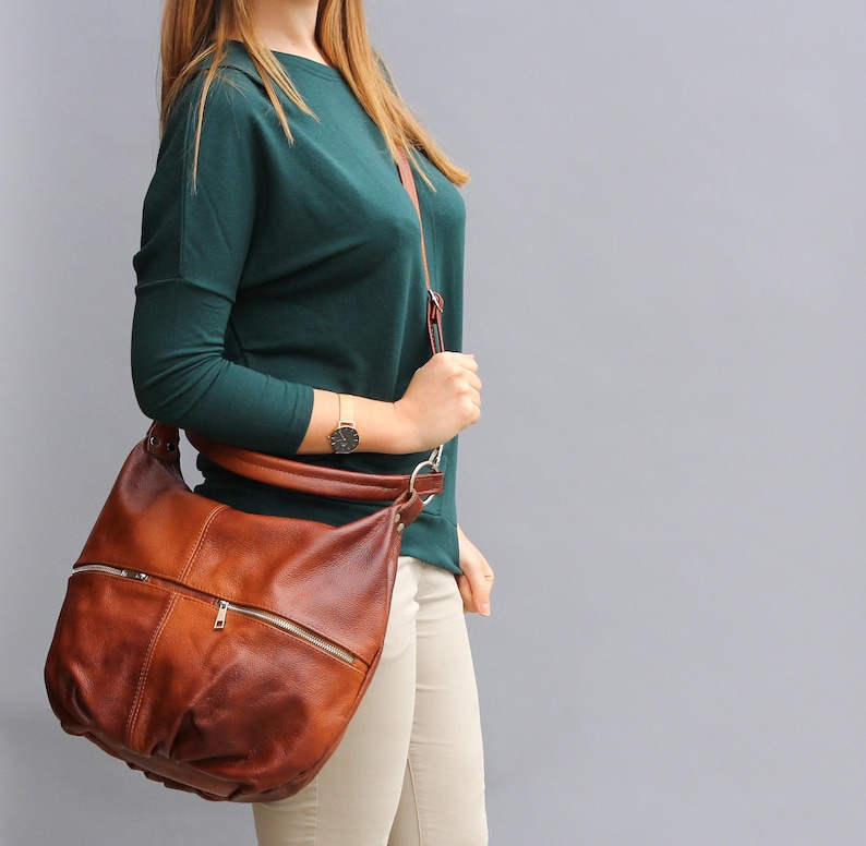 May include: A brown leather shoulder bag with a zippered pocket and a long strap. The bag is worn by a person wearing a teal sweater and beige pants.