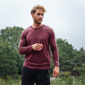 May include: A burgundy long-sleeve shirt with a crew neck. The shirt has a subtle heathered texture and a small logo on the left sleeve. The person is wearing black shorts. The background features green trees and a cloudy sky.