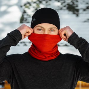 May include: A person wearing a black beanie with a white logo and a red neck gaiter. The person is pulling up the neck gaiter over their face. They are wearing a dark gray long-sleeve shirt.