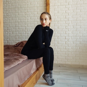 May include: A black long-sleeved thermal shirt with thumbholes and matching black thermal leggings. The shirt features a small white logo on the left wrist cuff. The set is worn with gray and black heathered knit socks.