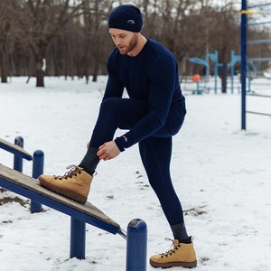 May include: A man wearing a navy blue long-sleeved shirt, navy blue thermal underwear, and a navy blue beanie is putting on a pair of tan leather boots. He is standing on a wooden platform with blue metal supports.