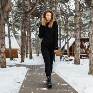 May include: A person wearing a black long-sleeved shirt and black leggings walks on a snow-covered path. The person is wearing black boots. The background includes trees, snow, and small wooden buildings.