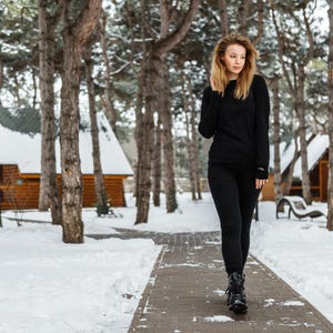 K&ouml;nnte beinhalten: Eine Person in einem schwarzen Langarmshirt und einer schwarzen Hose geht auf einem Steinweg in einem verschneiten Wald. Die Person tr&auml;gt schwarze Stiefel. Holzh&uuml;tten sind im Hintergrund zu sehen.