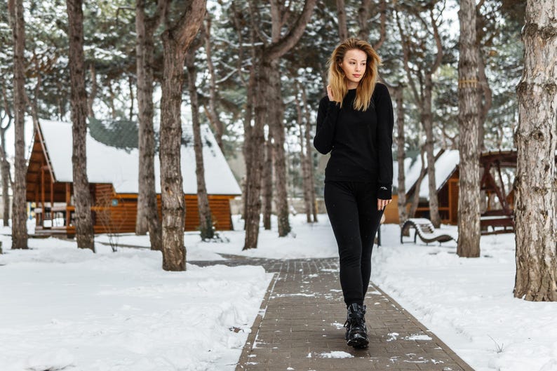 K&ouml;nnte beinhalten: Eine Person in einem schwarzen Langarmshirt und einer schwarzen Hose geht auf einem Steinweg in einem verschneiten Wald. Die Person tr&auml;gt schwarze Stiefel. Holzh&uuml;tten sind im Hintergrund zu sehen.