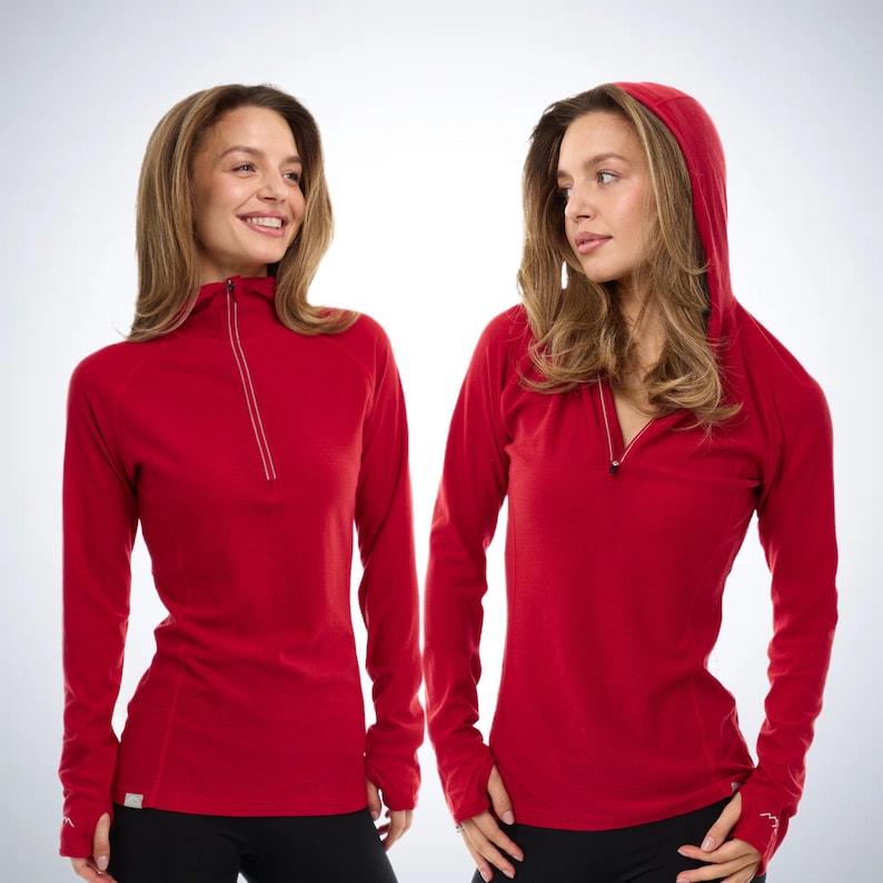 May include: Two long-sleeved red athletic shirts with a quarter-zip neckline. One shirt has a hood. Both shirts have thumbholes and a small logo on the sleeve. The shirts are paired with black athletic pants. The background is white.