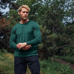 May include: A long-sleeved, forest green shirt with a crew neck. The shirt is paired with dark navy blue pants. The person is standing outdoors in a natural setting with trees and greenery in the background.