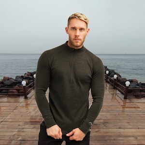 May include: A dark green long-sleeved shirt with a quarter-zip collar. The shirt has a slim fit and is made of a textured material. The person in the image is standing on a wooden dock near the ocean.