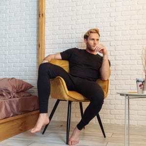 May include: A dark gray thermal shirt and matching pants. The outfit is worn by a person sitting in a yellow chair. The background includes a white brick wall, a bed with a brown pillow, and a small side table.