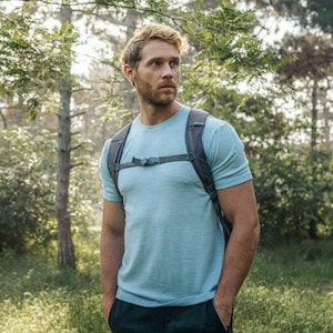 May include: A man wearing a light blue t-shirt with a gray backpack standing in a forest.