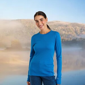 May include: A long-sleeved, crew neck shirt in a vibrant blue color. The shirt has a slim fit and is made of a soft, possibly thermal material. The model is smiling, and the background features a scenic landscape.