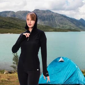 May include: Black hooded long-sleeve shirt with a quarter-zip closure. The shirt is paired with black leggings. The model is standing in front of a lake and mountains, with a blue tent in the background. The shirt has a small logo on the sleeve.