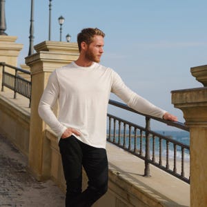 May include: A long-sleeved, off-white shirt and black pants. The shirt has a crew neck and a relaxed fit. The person is standing outdoors near a stone wall and a metal railing, with the ocean in the background.