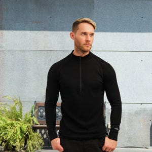 May include: Black long-sleeve shirt with a quarter-zip collar. The shirt has a slim fit and a small mountain logo on the left wrist. The model is wearing the shirt in front of a gray and blue wall.