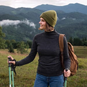 May include: An olive green knit beanie, a charcoal gray long-sleeve mock neck shirt, blue denim jeans, and a brown backpack. The gear includes two trekking poles with black handles and green shafts, held in a natural outdoor setting.