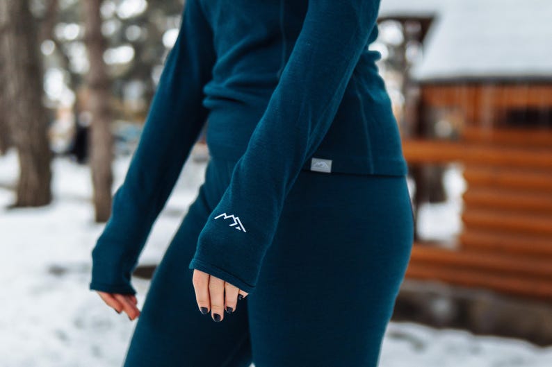 K&ouml;nnte beinhalten: T&uuml;rkisfarbenes Langarm-Top und Leggings-Set. Das &Auml;rmelb&uuml;ndchen hat eine wei&szlig;e Berg-Grafik. Das Set besteht aus einem weichen, m&ouml;glicherweise thermischen Material, das f&uuml;r kaltes Wetter geeignet ist. Das Outfit wird im Freien in einer verschneiten Umgebung gezeigt.