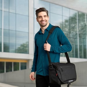 May include: A man wearing a teal button-down shirt and black pants, carrying a black messenger bag with a shoulder strap. The bag has a zippered pocket and a reflective strip. The background features a modern building with large windows.