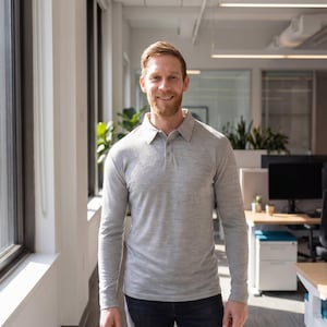 May include: A man wearing a light gray long-sleeve polo shirt with a buttoned collar. The shirt is made of a soft, textured fabric. The man is standing in an office setting with large windows and a desk in the background.