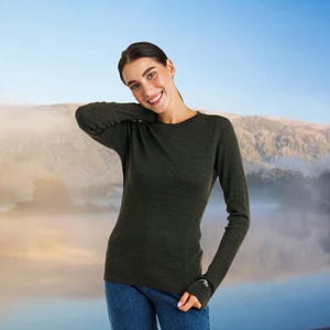 May include: A dark green long-sleeved shirt with a crew neck. The shirt is made of a soft material and is worn with blue jeans. The shirt has a small white logo on the left wrist. The background is a blurred landscape with a blue sky.