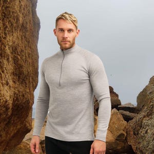 May include: A light gray long-sleeved shirt with a quarter-zip collar. The shirt is made of a soft, textured material. The model is wearing black pants. The background features rock formations and a cloudy sky.
